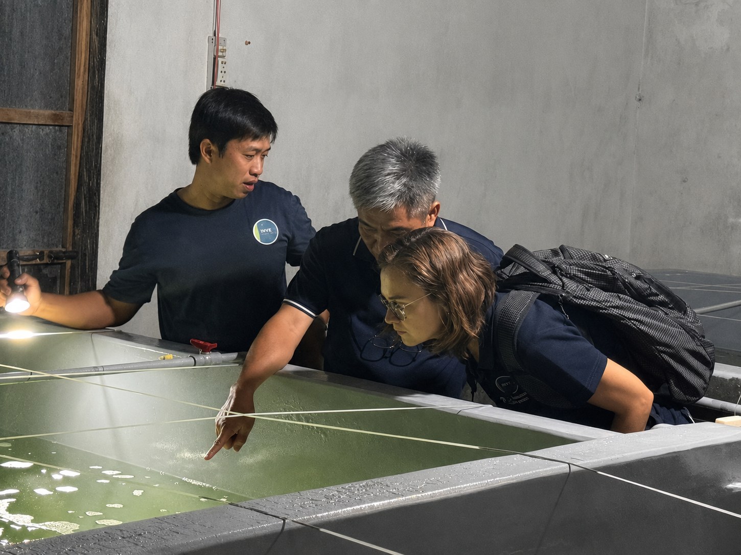 Técnicos de INVE Aquaculture y Eva Werbrouck observan un tanque de cultivo larvario de camarón con agua verde, iluminado con una linterna