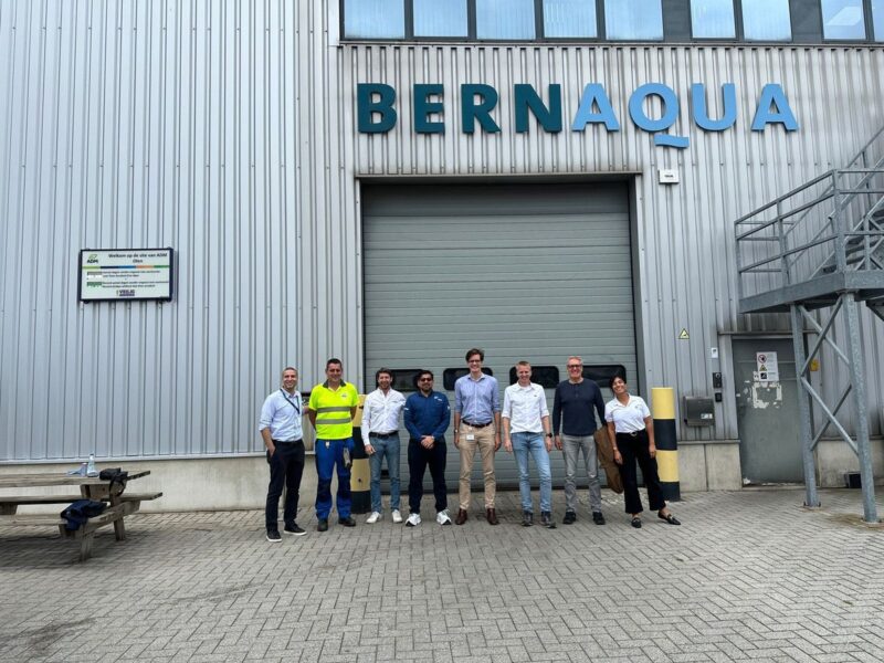 Ocho personas de pie frente a la fachada metálica de la planta Bernaqua, con el letrero de bienvenida al sitio de ADM en Olen, Bélgica